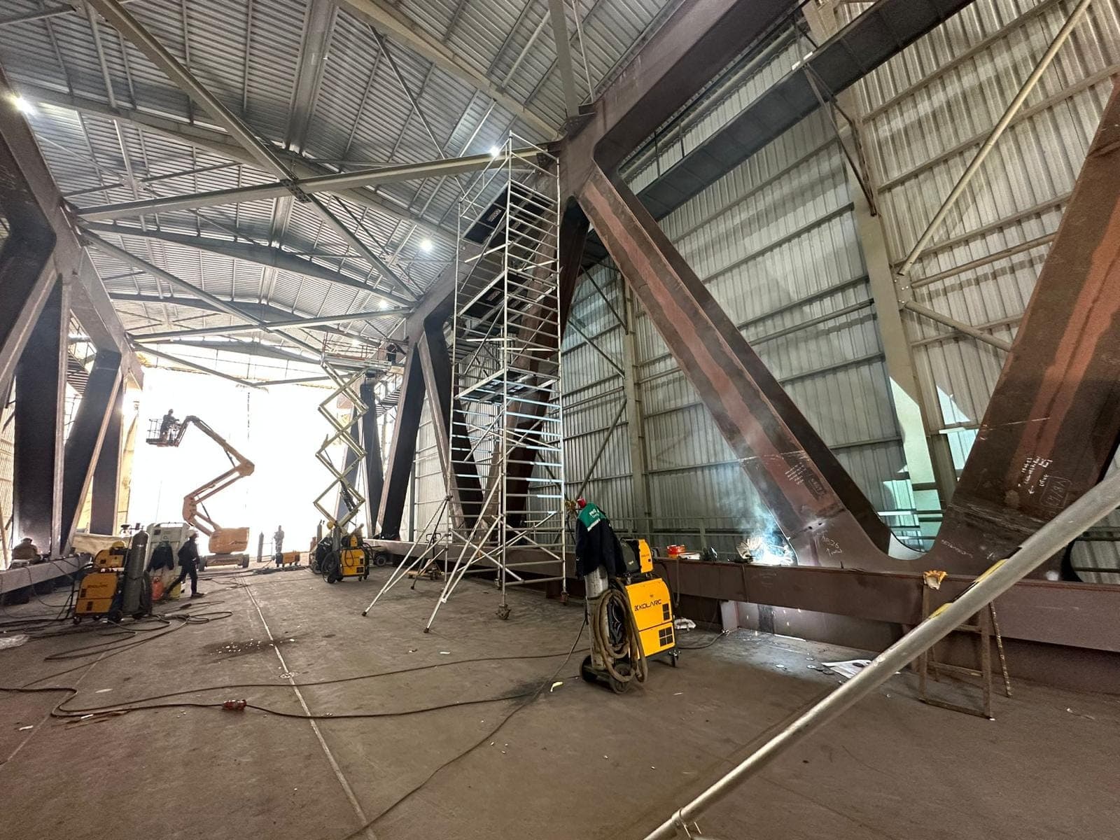 Kapalı imalat holünde kurulmakta olan çelik köprü; henüz boyanmamış kahverengi çapraz elemanlar, iskeleler, sepetli platform ve çalışan ekip