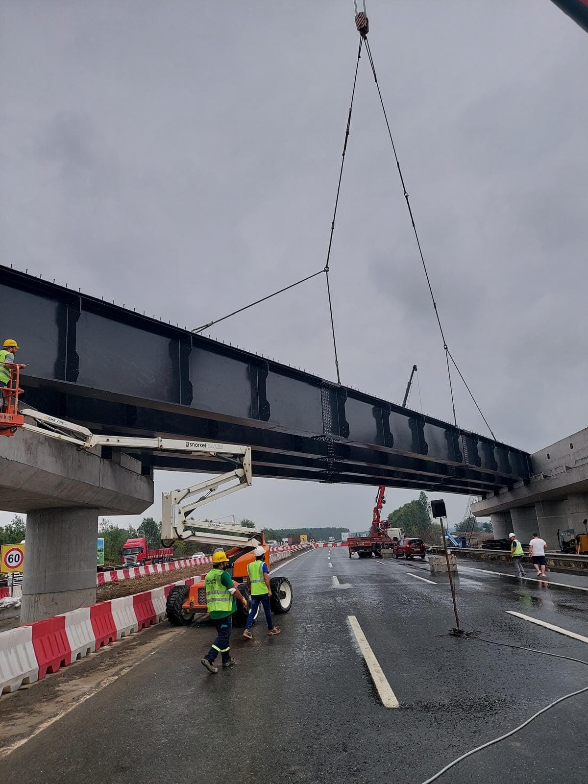 Yağmurlu havada otoyol üzerine halatlarla indirilen siyah boyalı çelik köprü kirişi; altta trafik bariyerleri, sepetli platform ve reflektörlü yelekli ekip