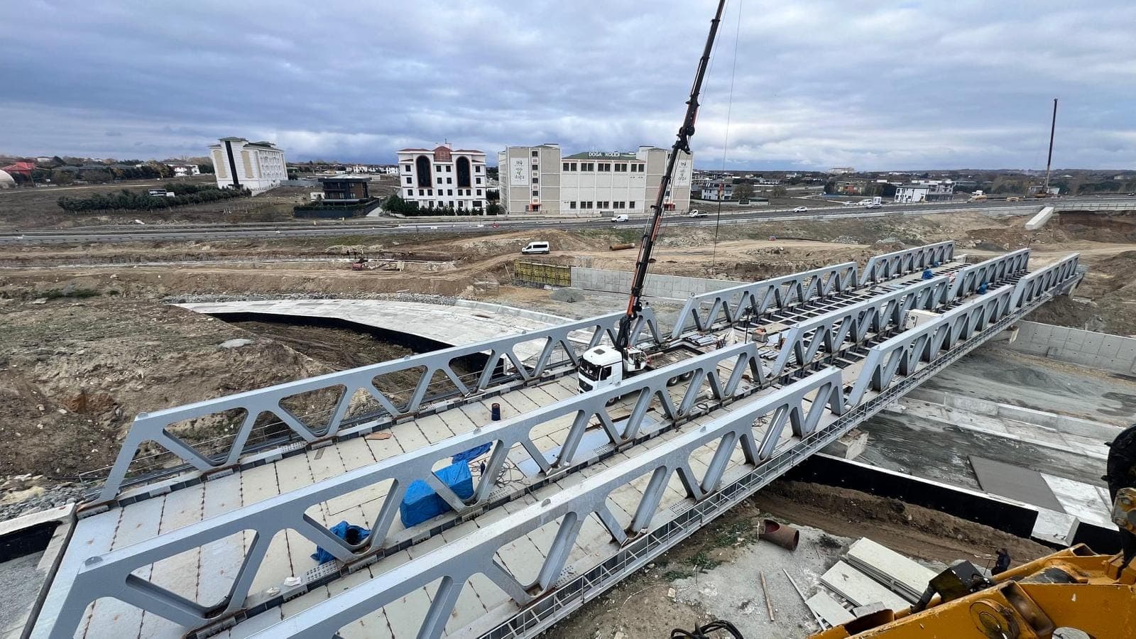 Gri boyalı iki kafes kiriş, bir su geçişi üzerindeki betonarme mesnetlere mobil vinçle indirilirken; arkada yerleşim ve yol, önde kazı yapılmış şev