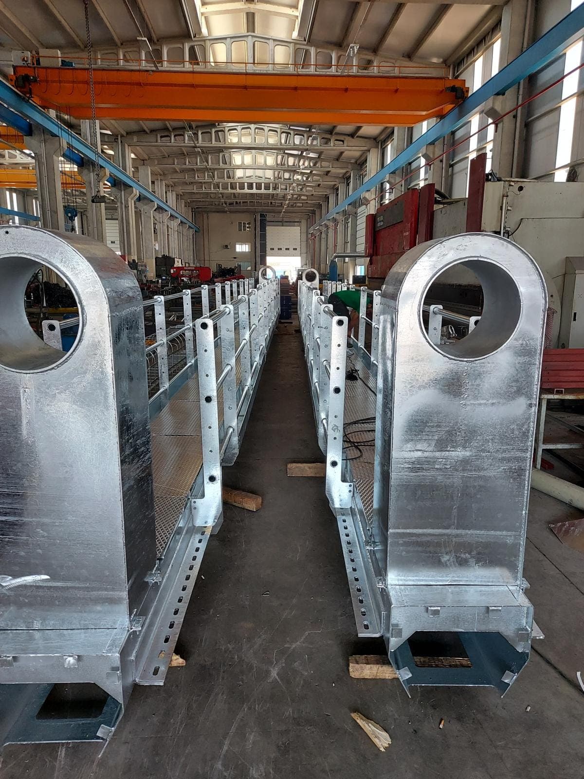 İmalat holünde yan yana duran iki sıcak daldırma galvanizli geçiş portalı; kutu kolonların üst ucunda ekipman için büyük dairesel açıklık, aralarında genişletilmiş metal döşemeli ve boru korkuluklu servis platformu, üstte turuncu gezer köprülü vinç