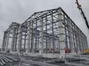 Hangarın köşeden görünümü; sıra sıra prefabrik beton kolonlar, üstlerinde çelik kafes makaslar, kolonlar arasında yatay çelik bağlantılar, zeminde istiflenmiş profiller