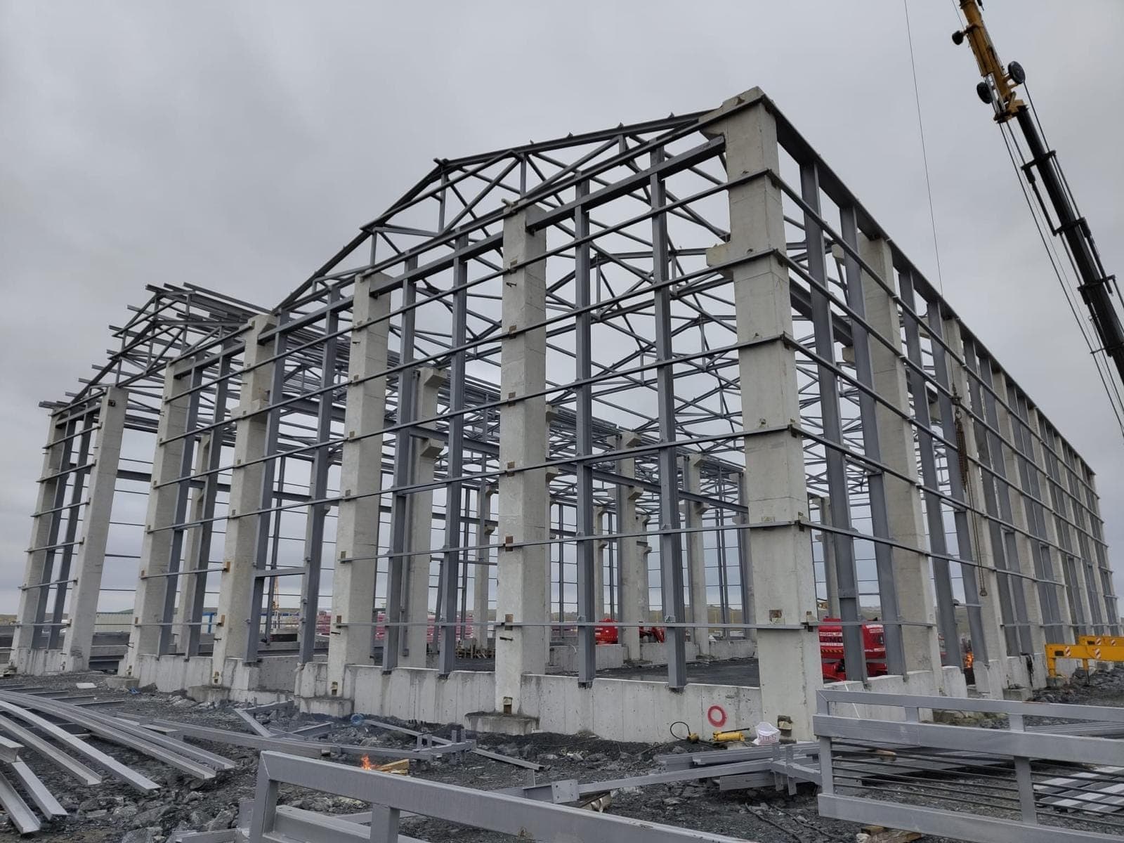 Hangarın köşeden görünümü; sıra sıra prefabrik beton kolonlar, üstlerinde çelik kafes makaslar, kolonlar arasında yatay çelik bağlantılar, zeminde istiflenmiş profiller