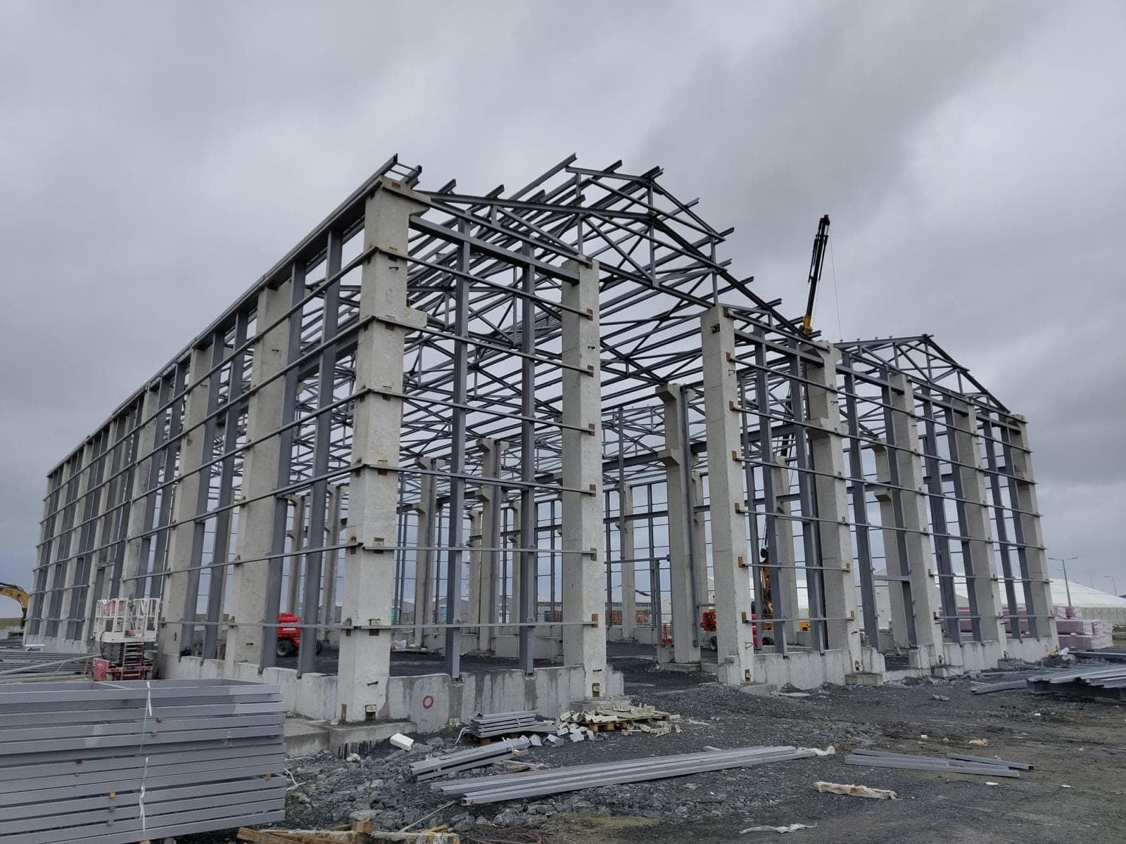 Hangarın yan cephesi; beton kolonlar arasındaki yatay bağlantılar ve çatı makaslarının eğimi görünüyor, zeminde montaj bekleyen çelik parçalar