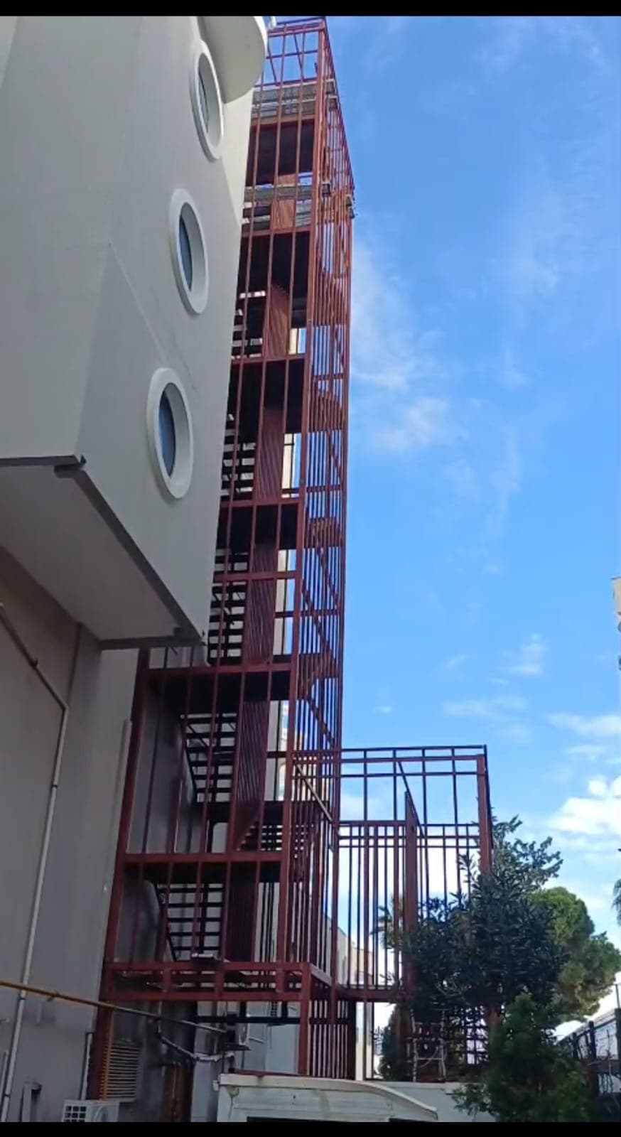 Bina cephesi boyunca yükselen, kırmızı boyalı dikey çubuk kaplamalı kule tipi çelik yangın merdiveni