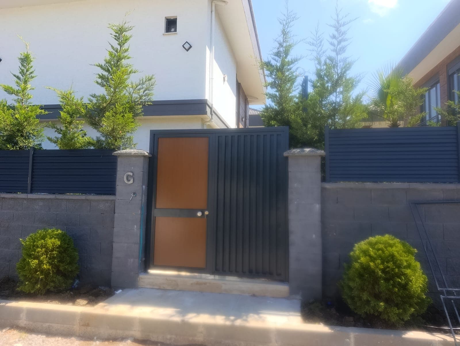 Antrasit renkli, ahşap görünümlü panelli yaya kapısı ve dikey lamelli kayar araç kapısı; iki yanında duvar üstü panjur çit korkuluğu